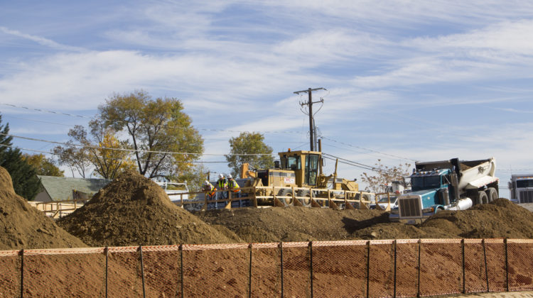 A yellow construction vehicle is sitting on top of a pile of dirt as construction works look down at the construction site