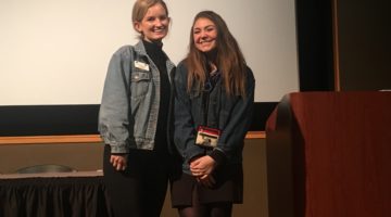 two-women-stand-on-stage-and-smile-at-camera-in-joe-crowley-student-union-theater
