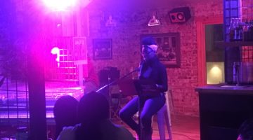 Girl sits on stool in front of an audience and reads poetry.