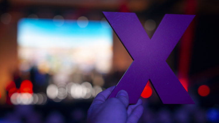 Someone holds up an X in front of the camera, while the stage at TEDx is blurred in the background.