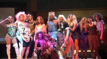 Group of drag queens, a drag king and some performers pose on stage for pictures together.