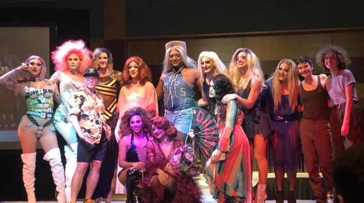 Group of drag queens, a drag king and some performers pose on stage for pictures together.