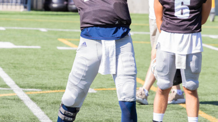 Redshirt freshman Carson Strong, wearing a number twelve practice jersey, throws a football during practice. Behind him is fellow quarterback Hamish McClure, looking at Strong while he prepares to throw the football.