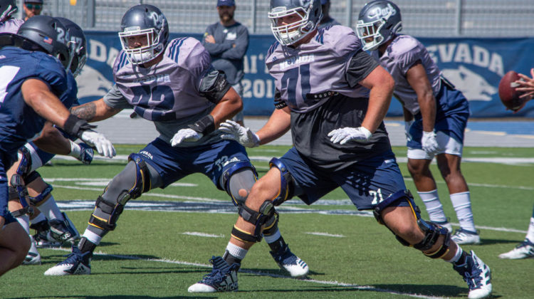 Wolf Pack linebacker Lucas Weber charges at two Nevada offensive linemen. Weber, wearing a dark blue jersey, goes against two linemen in off-pink jerseys.