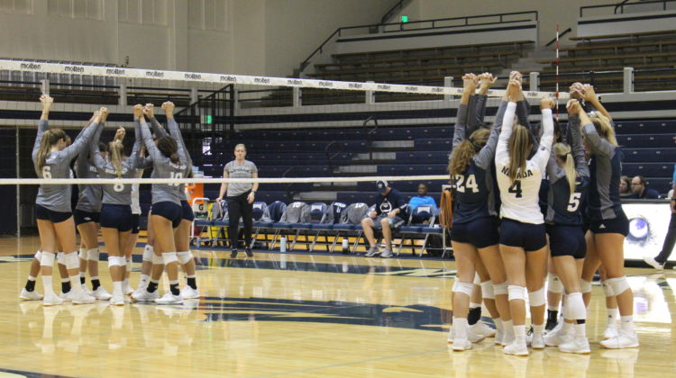 Nevada volleyball huddles in two groups on the court prior to opening of a scrimmage. On the far side of the court, half the team wears grey. On the closer side of the court, the team wears blue.