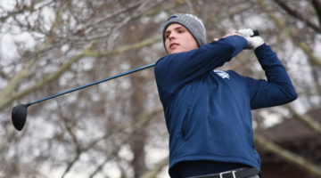 Nevada mens golfer Mitch Abbott swings his club during a round of golf. Abbott is wearing a dark navy blue jacket with the Nevada Wolf Pack emblem on the front left breast. He also wears a Nevada branded grey and white beanie on his head. Branches from tree can be seen overhead in the background.