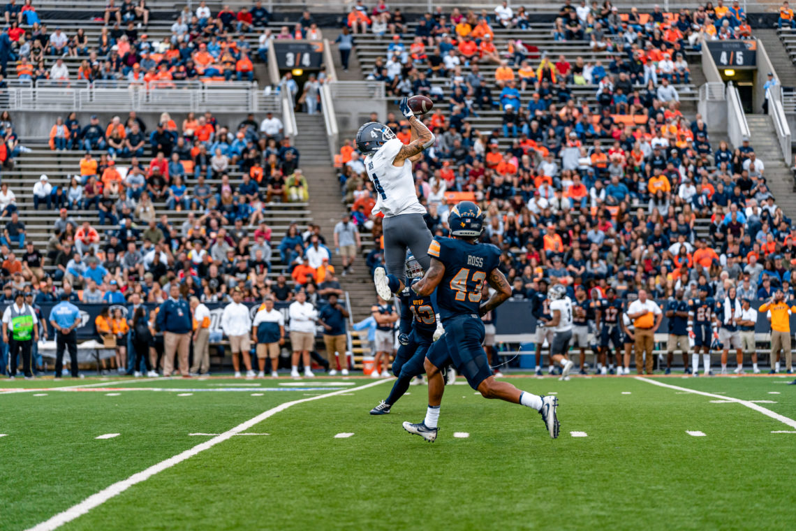 No. 4, Elijah Cooks, jumps over two UTEP defenders to secure a catch. Cooks is wearing grey pants and a white jersey. The two UTEP defenders are both wearing all blue uniforms with accents of orange.