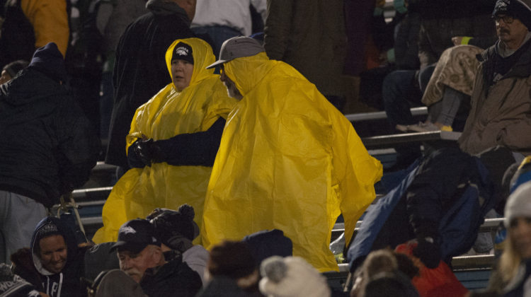 Two Nevada football sit in Mackay Stadium during a winter weather storm. The pair are wearing bright yellow ponchos with Nevada caps on their heads.