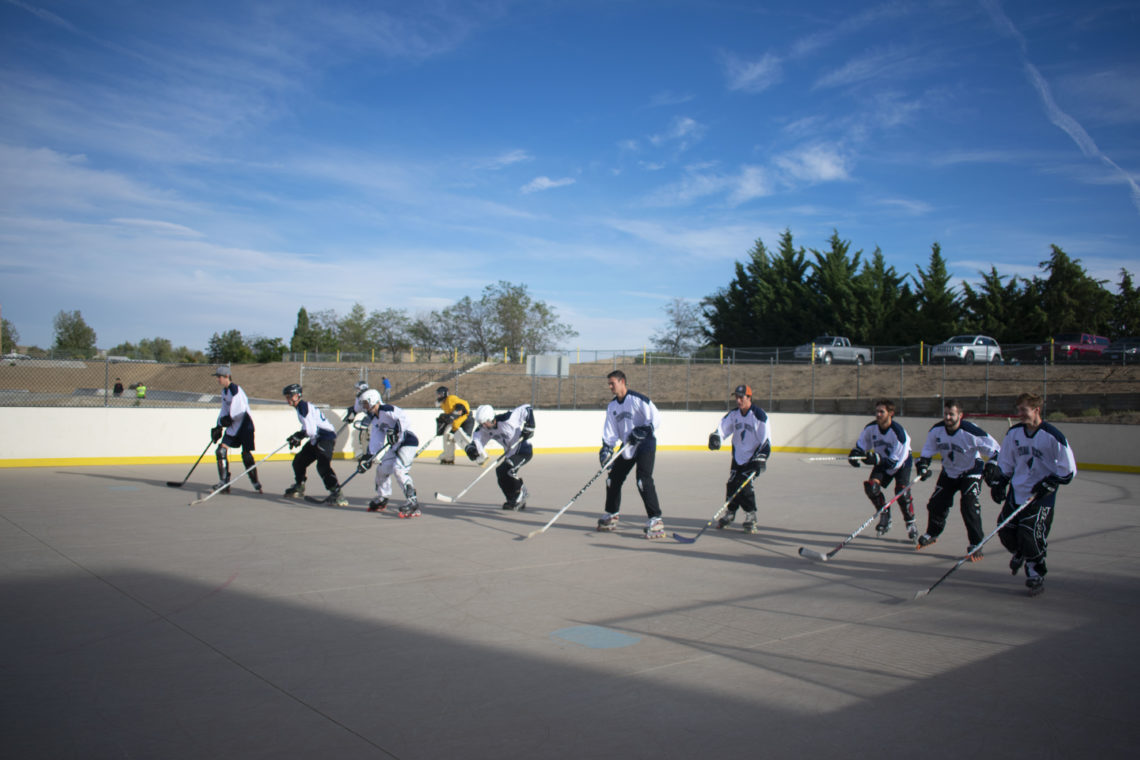 Nevada hockey players practice on a concrete court in the late afternoon. Each player is wearing a white jersey, expect for the goalie who is wearing white.