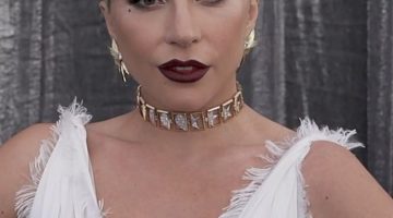 A close-up picture of Lady Gaga at a red carpet event. She has a white dress on with a gold choker. Her hair is blonde and slicked back.