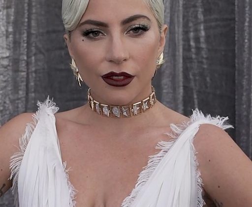 A close-up picture of Lady Gaga at a red carpet event. She has a white dress on with a gold choker. Her hair is blonde and slicked back.