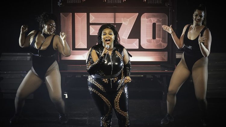 Three women in black leotards performing on a stage with the words "Lizzo" in pink behind them.