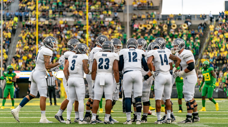 Nevada's offense, led by quarterback Carson Strong, huddles before a play against Oregon in Eugene, Oregon. Nevada is wearing all white, apart from their grey helmets.