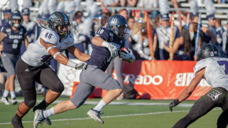 Ben Putman, wearing a No. 10 Blue Nevada jersey, runs between two Weber St. defenders. The two defenders are both wearing white jerseys with black pants.