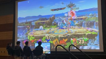 Four people sitting on stage playing a video game in front of a large projector. The video game on the projector shows an island and has four characters fighting.