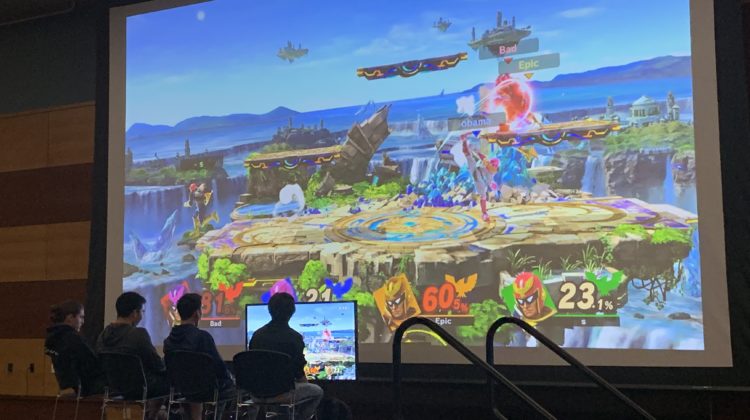 Four people sitting on stage playing a video game in front of a large projector. The video game on the projector shows an island and has four characters fighting.