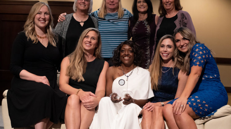 Nine women pose infront of a couch for a group photo. Five are sitting on the couch, and the other four are standing behind the couch.