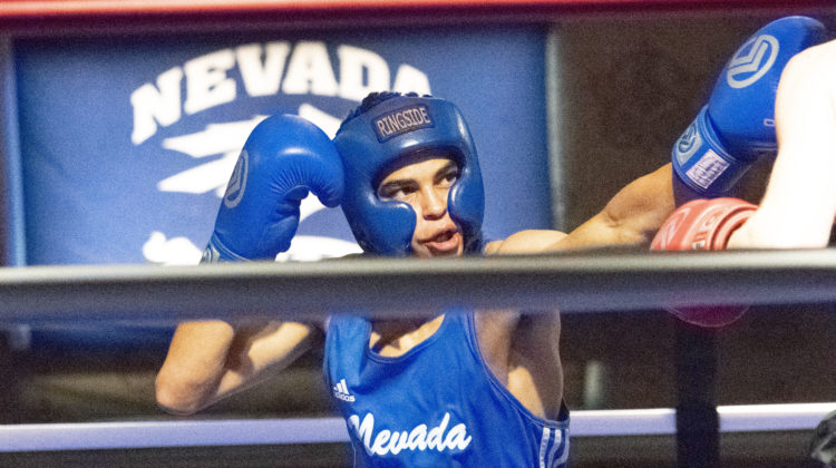 A Nevada boxer lunges at his opponent during a fight on Oct. 18, 2019. He is wearing a blue top with Nevada in white cursive writing on it. Also on his person, is blue boxing gloves and a blue boxing helmet. Behind him is a flag with the Nevada razor-wolf logo on it.