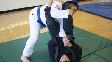 Two members of the UNR Brazilian Jiu-Jitsu practice holds on each other. One is wearing a white suit with a blue belt, while the other is wearing a black suit with the same colored belt.