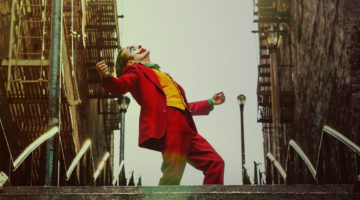 A man in a red suit with blue hair and white clown makeup stands on an outdoor staircase. The words "Joker" are in all caps on the bottom of the poster.