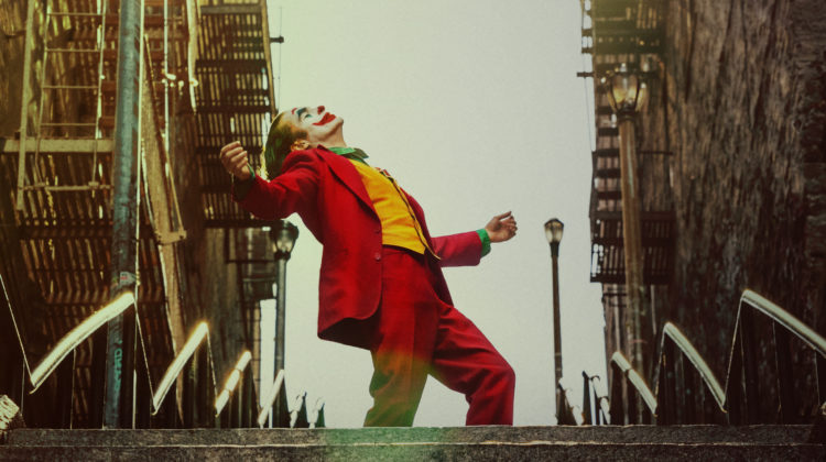 A man in a red suit with blue hair and white clown makeup stands on an outdoor staircase. The words "Joker" are in all caps on the bottom of the poster.