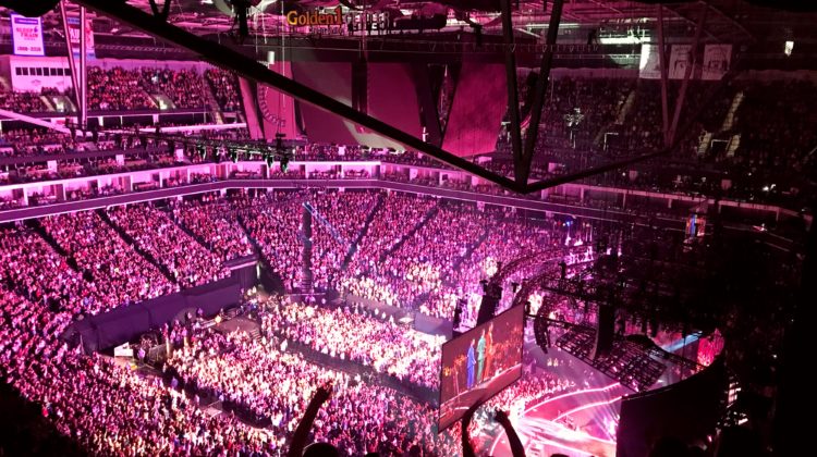 An area full of people at a Jonas Brothers concert. There is a screen that shows Joe Jonas, Nick Jonas and Kevin Jonas in blue, green and red suits. The lighting is all pink and the Golden1 Center logo is hung from the top of the arena.