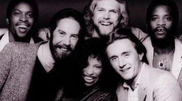 Pictured in black and white, five men in suits are crowded around Chaka Khan. All are smiling at the camera.