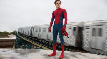 Tom Holland in a Spider-Man costume, which is red and blue with a black spider design in the middle. He is standing on a white moving train.