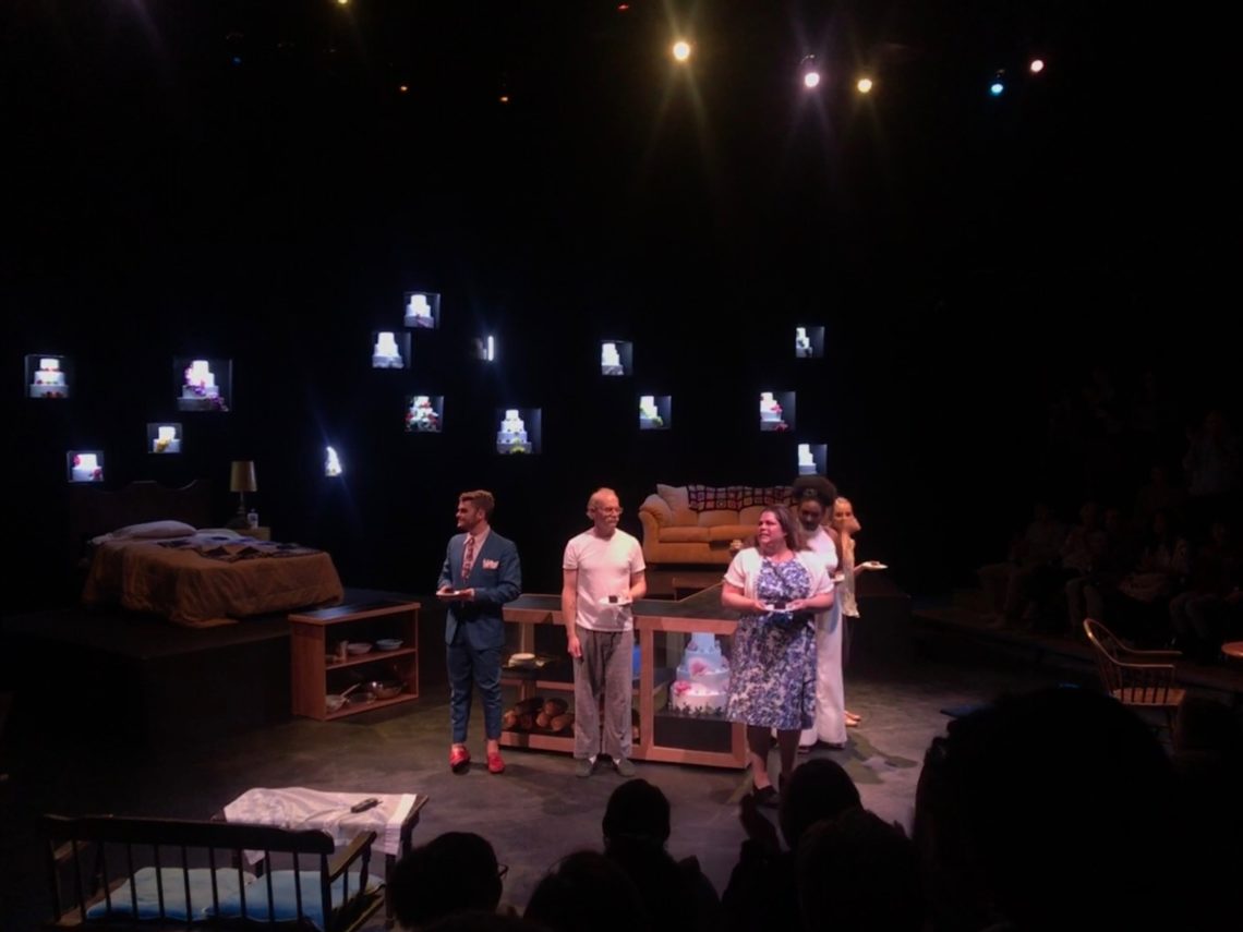 This picture is placed in a theater setting with a bed, a couch and a table. Four people—one man in a blue suit, one man in a white t-shirt and sweatpants, a woman in a blue flowered dress and a woman with a long white dress—are in the spotlight. There are light up cases with cakes in them in the background.