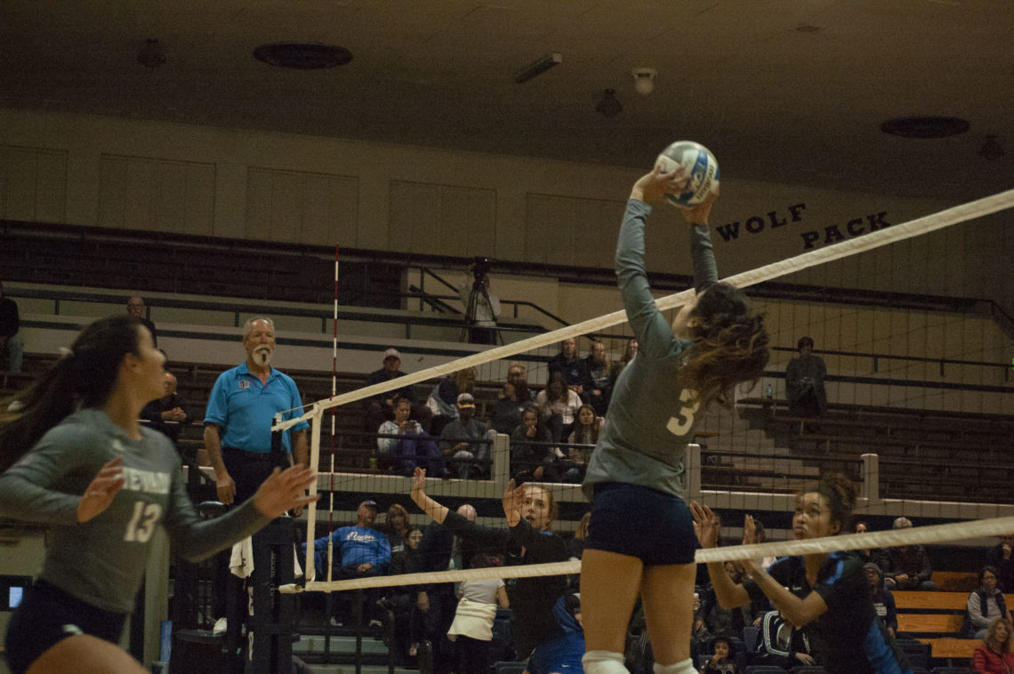 Dalyn Burns leaps in front of the net, towards a midair volleyball. Burns is wearing a long sleeve grey shirt with blue shirts and long white socks.