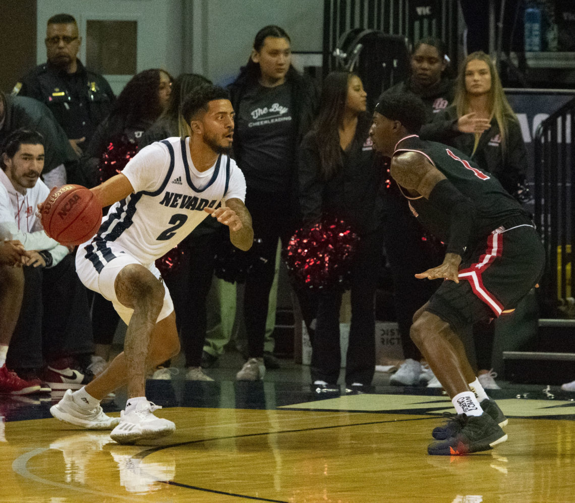 Jalen Harris goes on-on-one against a Cal State East Bay player. Harris is wearing all white, with blue accents. He is holding a basketball in his right hand.