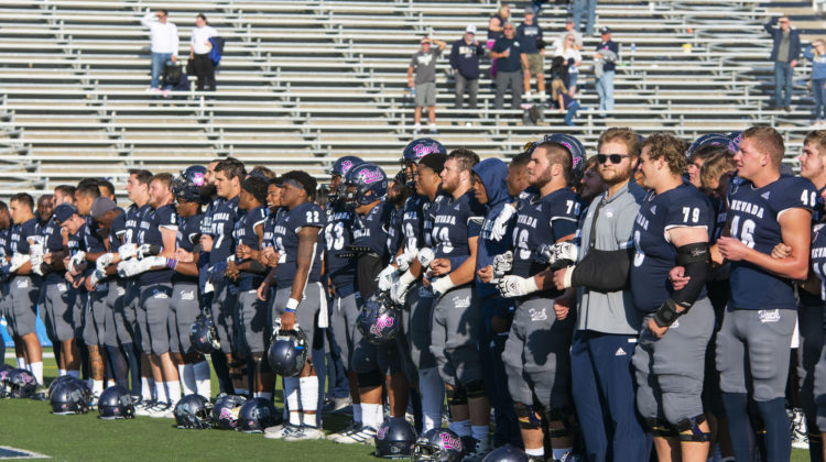 Nevada Wolf Pack football players stand side by side with their arms locked. All are wearing blue and silver uniforms. Some are wearing their helmets that have the word 'Pack' in pick lettering on them.