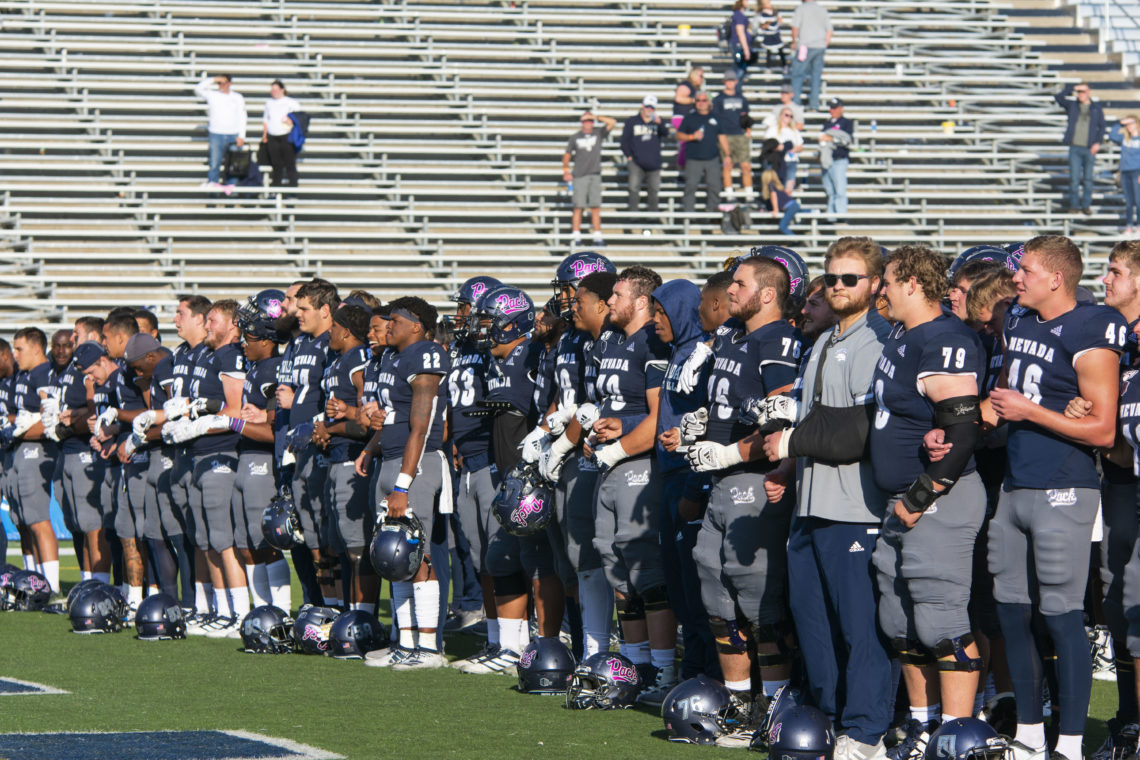 Nevada Wolf Pack football players stand side by side with their arms locked. All are wearing blue and silver uniforms. Some are wearing their helmets that have the word 'Pack' in pick lettering on them.