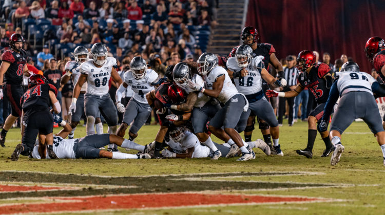 Multiple members of the Nevada football team pile onto a San Diego State running back. Nevada is wearing white jerseys with grey pants.