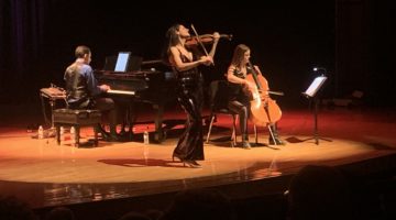 Take3 performing on stage. One woman is playing the violin, another is playing the cello and one man is playing piano.