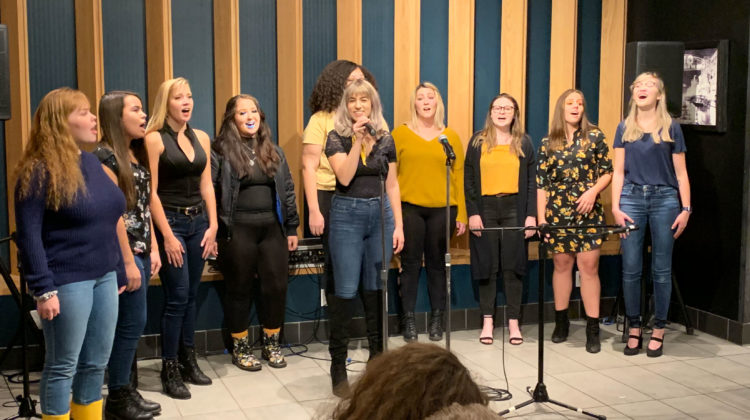 An a capella group of 10 people perform at the Joe Crowley Student Union Starbucks. All ten girls are singing in front of tan colored banners.