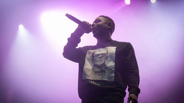 Kendrick Lamar performing at a festival. Lamar is wearing a black sweatshirt with Frankenstein on it and is holding a mic up in front of purple strobe lights.