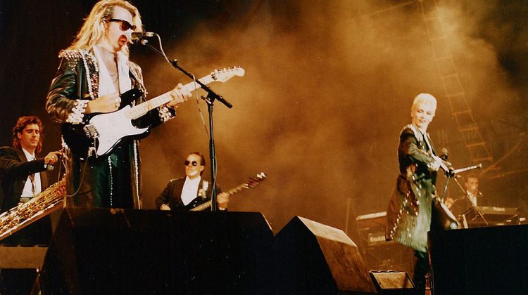 Annie Lennox and Dave Stewart (Eurythmics) performing. Stewart is playing a guitar and Lennox is dancing in a leather coat.
