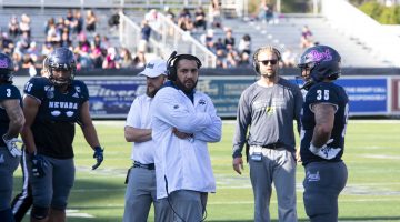 Coach Vai Taua coaches his younger brother on the sideline of Mackay Stadium. Taua is wearing a white windbreaker with headset fixed to his head.
