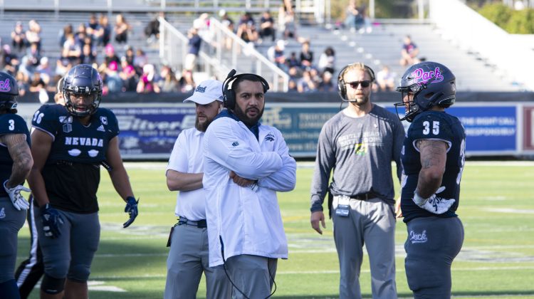 Coach Vai Taua coaches his younger brother on the sideline of Mackay Stadium. Taua is wearing a white windbreaker with headset fixed to his head.