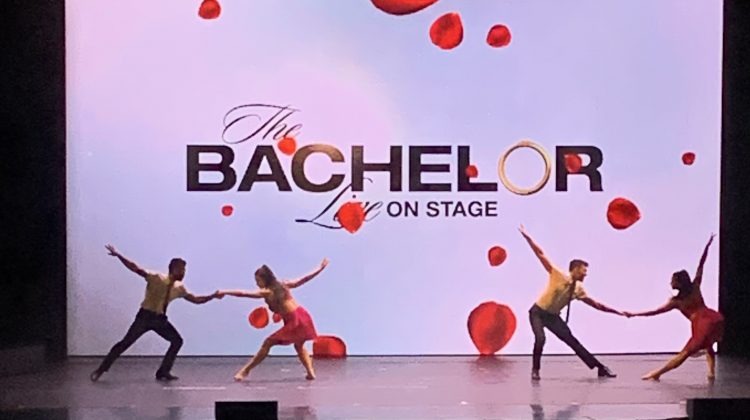 Four people dancing on stage with a white backdrop that says "The Bachelor Live On Stage."