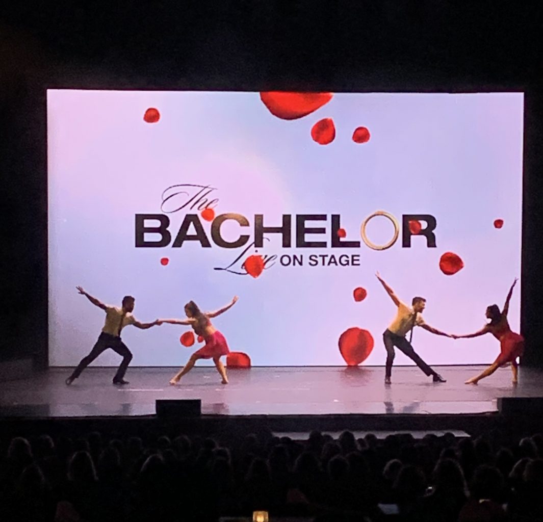 Four people dancing on stage with a white backdrop that says "The Bachelor Live On Stage."