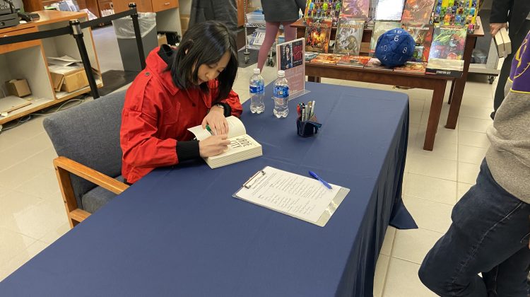 Novelist Fonda Lee signing a book on a blue table wearing a red coat.