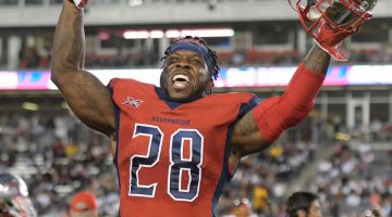 James Butler celebrates with his arms raised following a victory over the Los Angeles Wildcats. Butler is wearing a red jersey with the No. 28 in blue and white on his chest. In his left hand, he holds his silver football helmet.