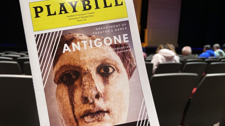 A close-up of a Playbill booklet with the ancient Greek statue on the cover. The Playbill says "Antigone" in bold white letters and also includes dates on the bottom half. An audience and a theater stage are also pictured behind the Playbill, but out of focus.