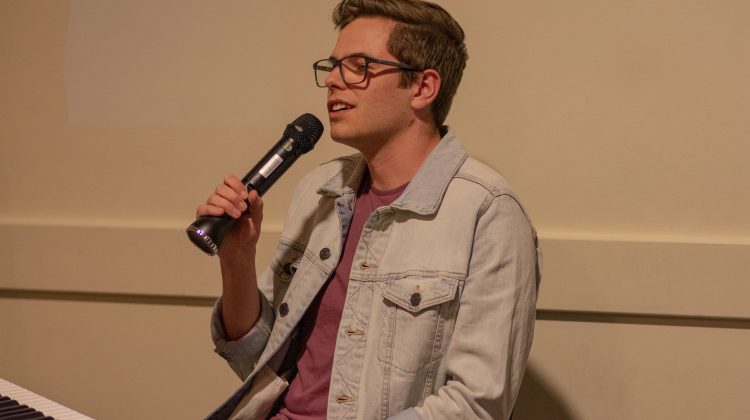 Ben Torvinen, wearing glasses and a light washed denim jacket, sitting down by a keyboard and holding a microphone.