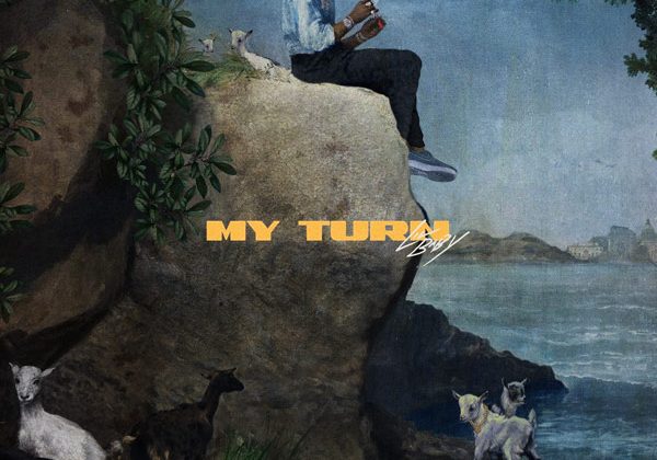 A man sits on a big rock with jeans and a sweatshirt on with the ocean behind him. The words "My Turn" are spread across the middle of the picture with yellow letters.