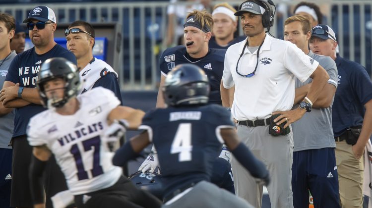 Jay Norvell watches from the sideline during a football game. Norvell is wearing grey pants and a white polo. In the foreground of the image, two football players run in front of him.