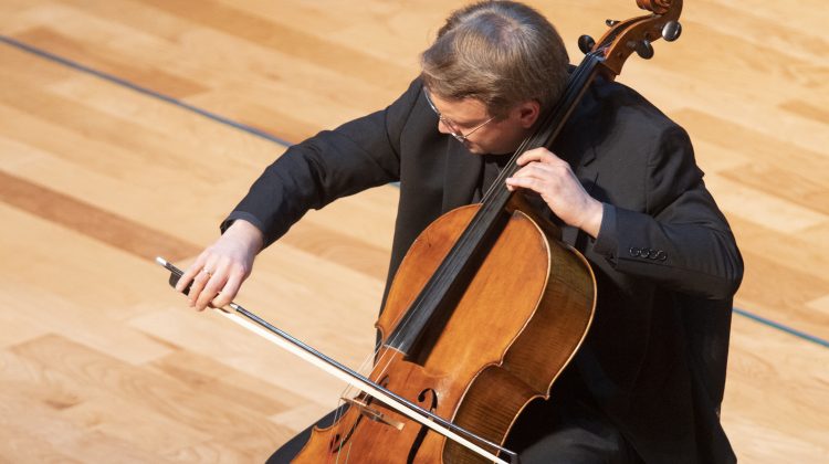 Man plays cello with the background of wood walls in a recital hall.