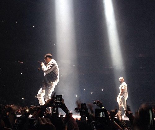 Drake and Kanye under spotlights on a black stage with audience in the background whilst they're wearing all white.